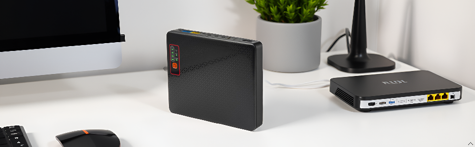 Black rectangular electronic device with textured surface and red LED indicators, placed on a white desk next to a plant, monitor, and network router.