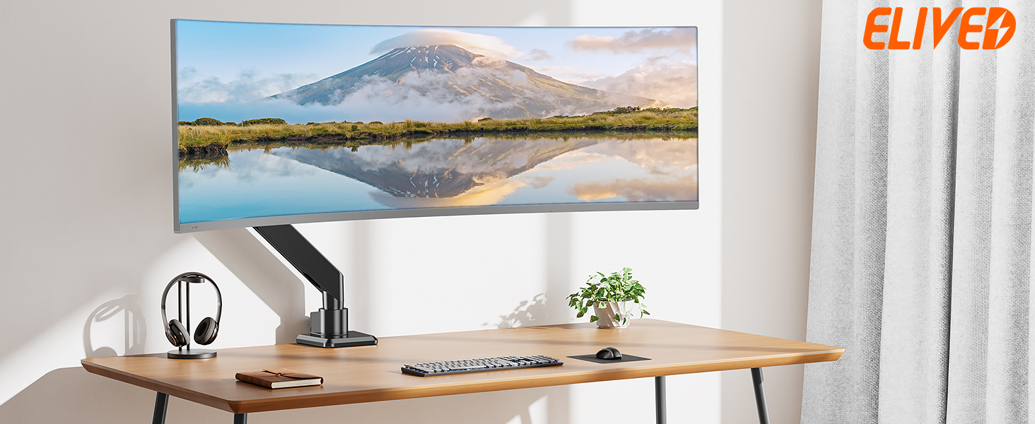 A modern workspace with a wooden desk featuring a mounted ultrawide monitor displaying a landscape image. The desk also holds a keyboard, headphones, and a small plant.