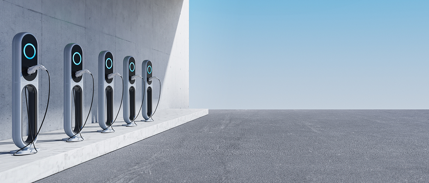 Row of black electric vehicle charging stations mounted along a white wall, extending into the distance against a blue sky.