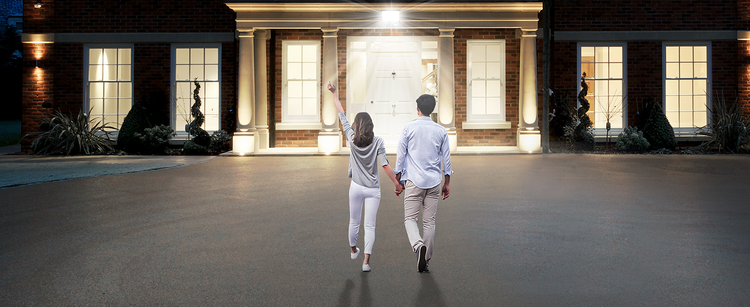 Illuminated entrance of a large house at night, with columns and multiple windows. Two figures walking towards the front door.