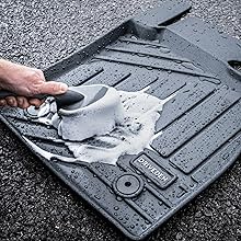 Black rubber car floor mat being cleaned with soap and water, showing deep grooves and channels designed for catching dirt and debris.