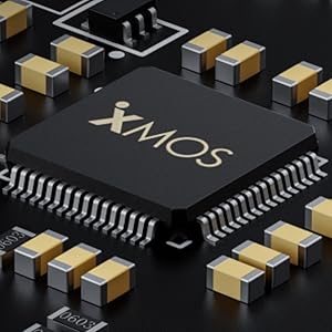 Text reads 'XMOS' on black microchip surrounded by gold-colored capacitors and electronic components on circuit board. Macro photography showing technical detail.