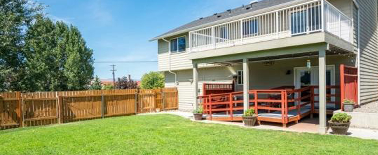 Two-story house with white siding, upper deck with white railing, and red wooden deck below, surrounded by fenced yard with green lawn.