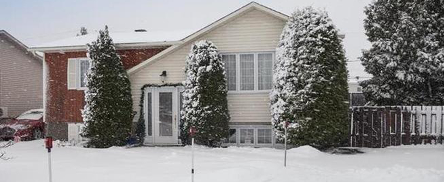 Single-story residential house with white siding covered in snow, featuring evergreen trees and a visible attached garage during winter.