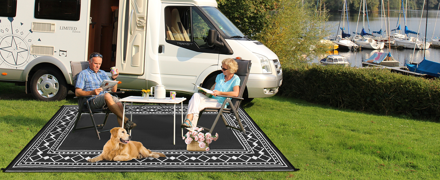 Outdoor camping scene with recreational vehicles in background. Large patterned outdoor mat on grass with dog sitting on it. Marina visible in distant background.