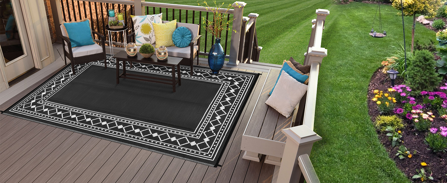 Outdoor deck scene with black and white patterned area rug, two blue chairs, and decorative garden with flowers and lawn in background.