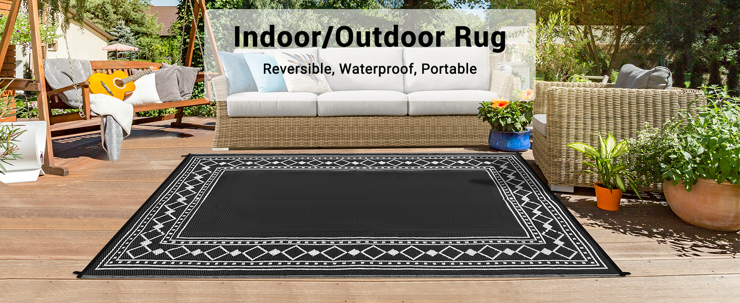Text reads 'Indoor/Outdoor Rug'. Large black rectangular area rug with ornate border pattern displayed on patio setting with lattice fence and potted plants.