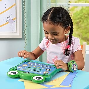 Young child playing with green educational tablet device on blue table. Device features colorful buttons, interactive screen display, and hand controls. Learning toy demonstrating interactive educational play.