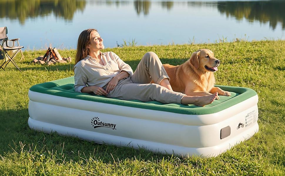 A raised air mattress in cream and green colors set up outdoors by a lake, with a person relaxing on it alongside a golden-colored dog.