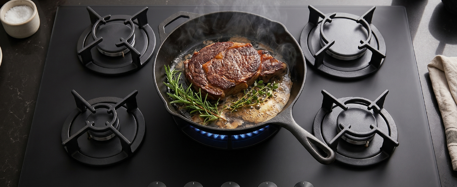 Overhead view of a black stovetop with four gas burners and a large pan containing seared meat with herbs and vegetables, professional kitchen setup.