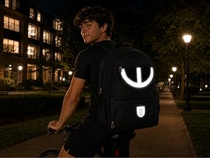 Black backpack with front zippered compartments and mesh pockets, hanging on chain-link fence with sports field background.