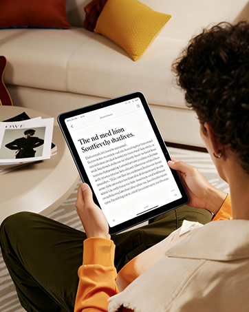 Person in blue clothing lying on bed using light blue tablet device, with white quilted pillow and wooden shelving with books and items visible in background.