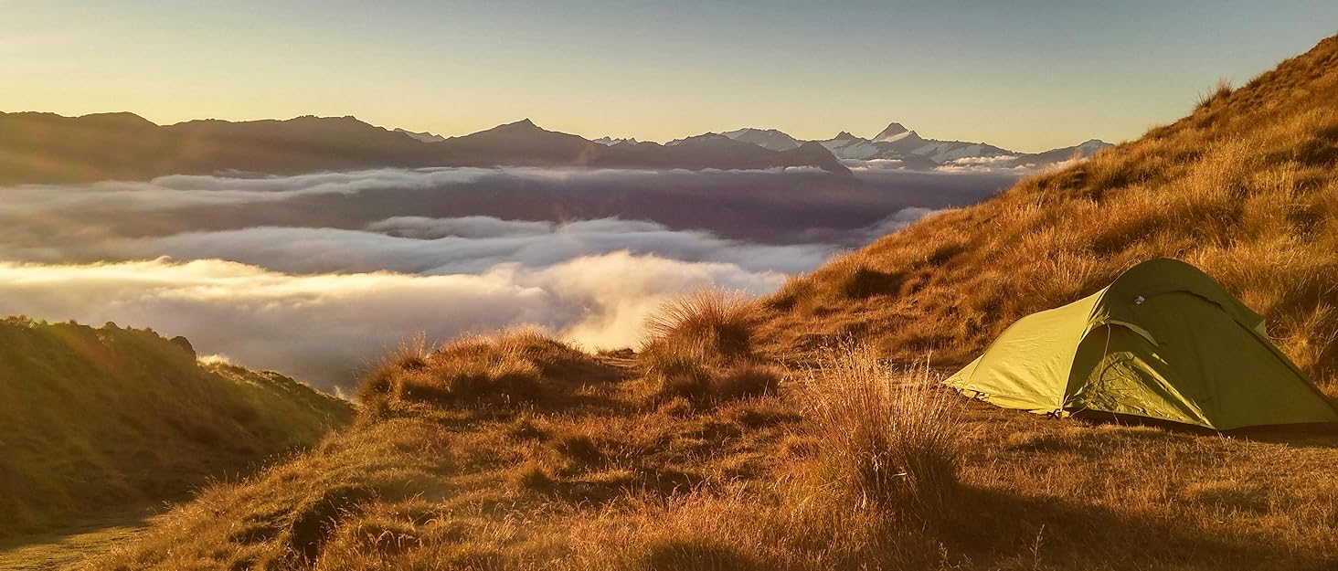 Mountain landscape photograph featuring golden grassland, misty valley, distant peaks, and yellow tent camping setup in natural outdoor wilderness setting.