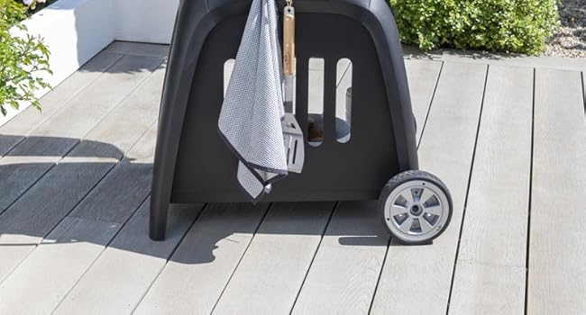 Outdoor charcoal kettle grill with dome cover, two-wheel base, mounted on wooden patio deck. Black metal construction with temperature gauge, cooking grate, and ash collection system visible. Green foliage backdrop with food items nearby.