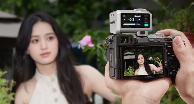 Digital camera with flip-out screen displaying image preview. Top-mounted accessory shows numerical readout. Camera held in hand, capturing outdoor portrait.