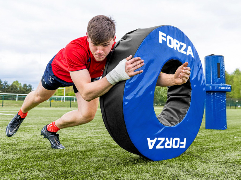 Text reads 'FORZA YZFOCP.' Person in athletic gear performing a horizontal plank exercise through a blue and black tire obstacle on grass field.