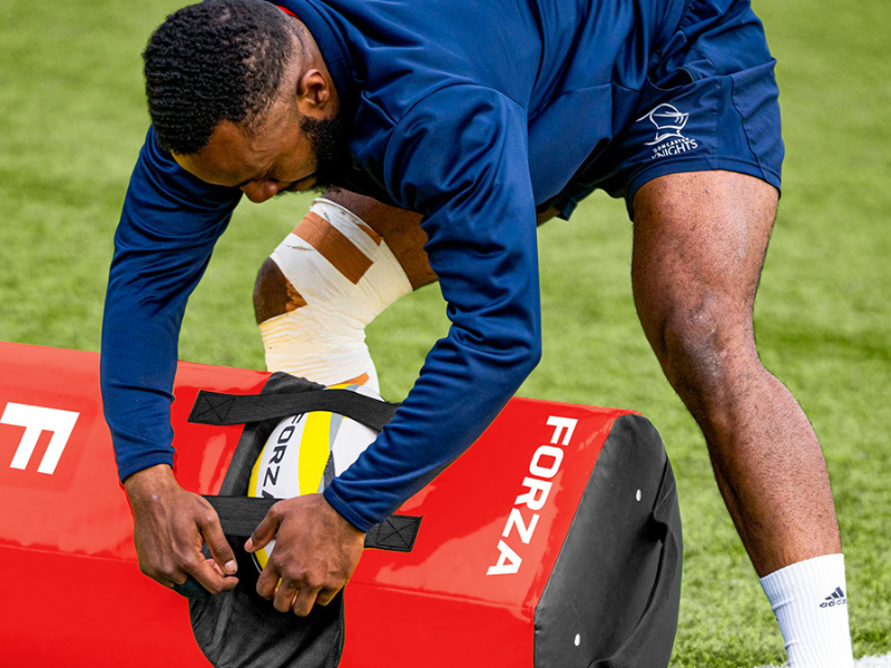 Person wearing blue jacket and white athletic tape wrapping on wrists, holding white and brown football/rugby ball on red equipment bag outdoors.