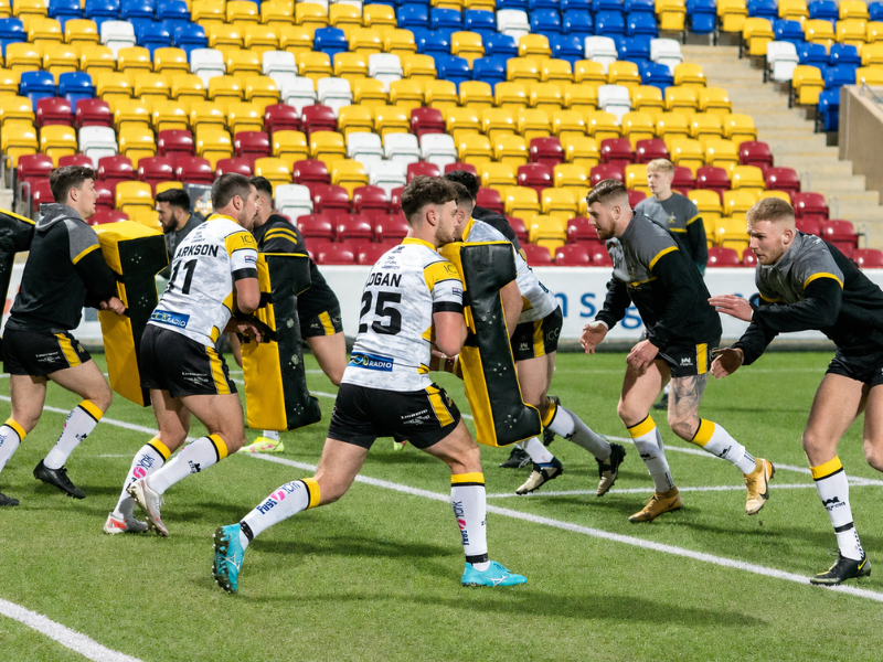 Professional lacrosse game in progress on field with colorful yellow, blue, and red stadium seating visible behind players. Multiple athletes in black and white uniforms engaged in competitive play with lacrosse sticks.