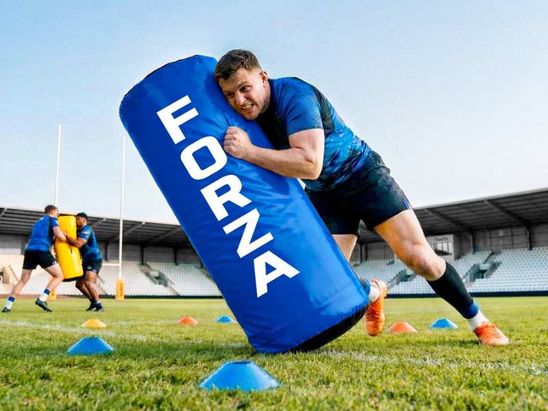 Text reads 'FORNY'. Male athlete in blue shirt pushing large blue foam blocking pad during outdoor rugby or football training on grass field with field markings visible.