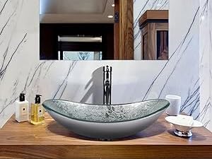Modern bathroom vanity with white countertop and gray oval vessel sink. Chrome faucet, gray tile wall, and mirror visible. White towels and plant accessories included.