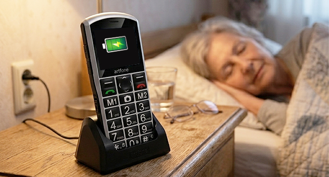 Black mobile phone charging on nightstand next to sleeping person in bed during evening. Device displays green battery indicator light while plugged into charging dock.