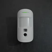Square white device with rounded corners mounted on wooden paneled wall. Device has 'AJAX' printed on front. Likely a smart home or security system component.