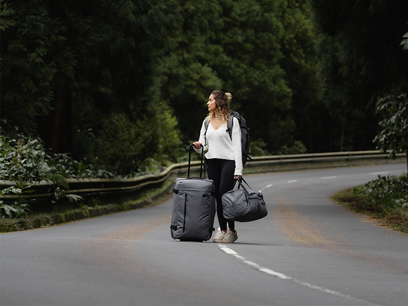 Woman in white shirt standing outdoors on road with black rolling luggage suitcase and matching black shoulder bag, demonstrating travel use.