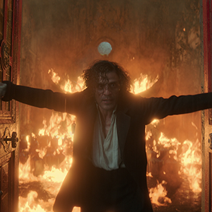 Silhouetted figure with arms outstretched standing in front of warm glowing lights and fire in dark atmospheric setting.