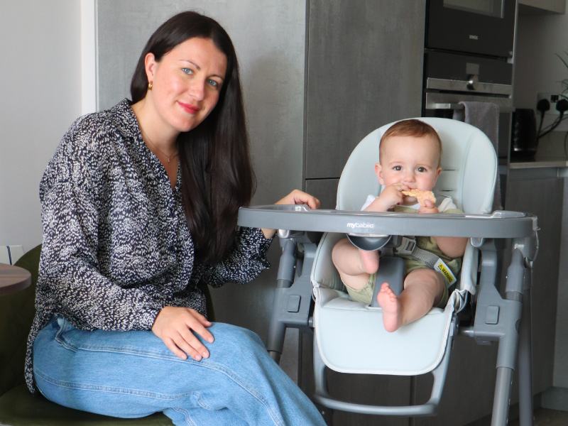Interior scene showing grey highchair with baby seated in it, accompanied by adult in patterned shirt and jeans.