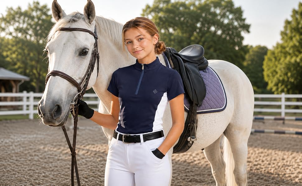 Woman in dark polo shirt and white riding pants standing beside white horse with halter in outdoor equestrian arena.