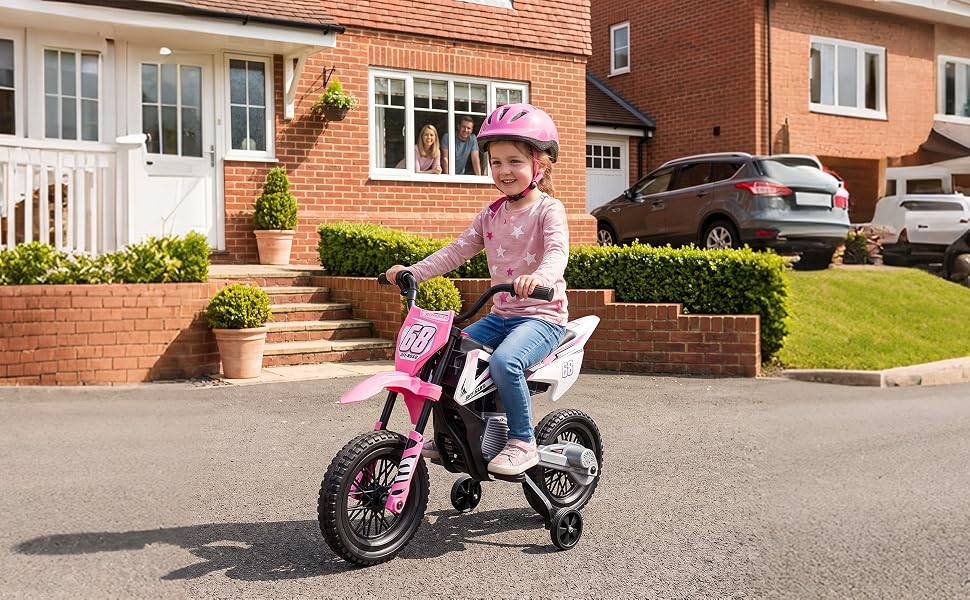 Child in pink helmet riding pink and black pedal bike on suburban driveway in front of brick house with landscaping, demonstrating balance bike usage outdoors.