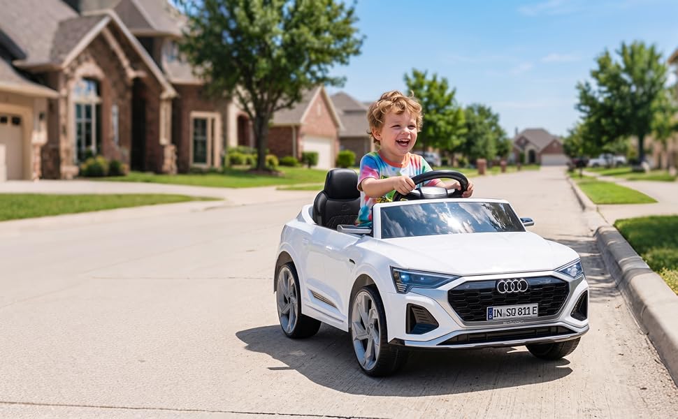 White ride-on toy car with children sitting on top, parked in residential driveway on sunny day with brick house and trees visible.