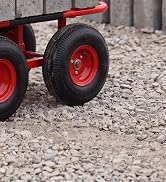 Side view of red and black pedal-powered ride-on toy with dual black tires and red metal wheel hubs on gravel ground.