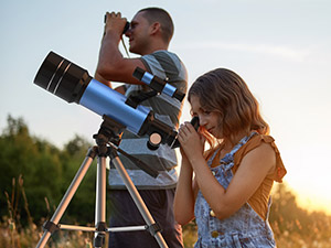 Two children outdoors during golden hour using telescope mounted on yellow tripod, looking through eyepiece at sky with grass field background.