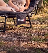 Black portable folding camping bench or stool with metal frame legs, positioned on outdoor ground with grass and natural terrain visible.