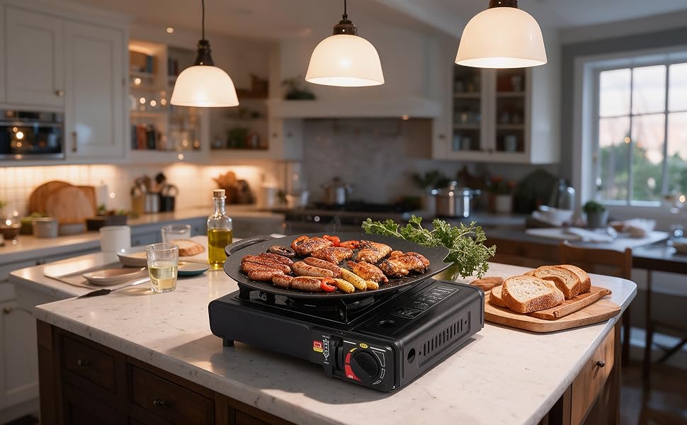 Portable black grill or griddle cooktop displayed on a kitchen island with food cooking on surface, surrounded by dishes and kitchen setting with pendant lights and shelving visible.